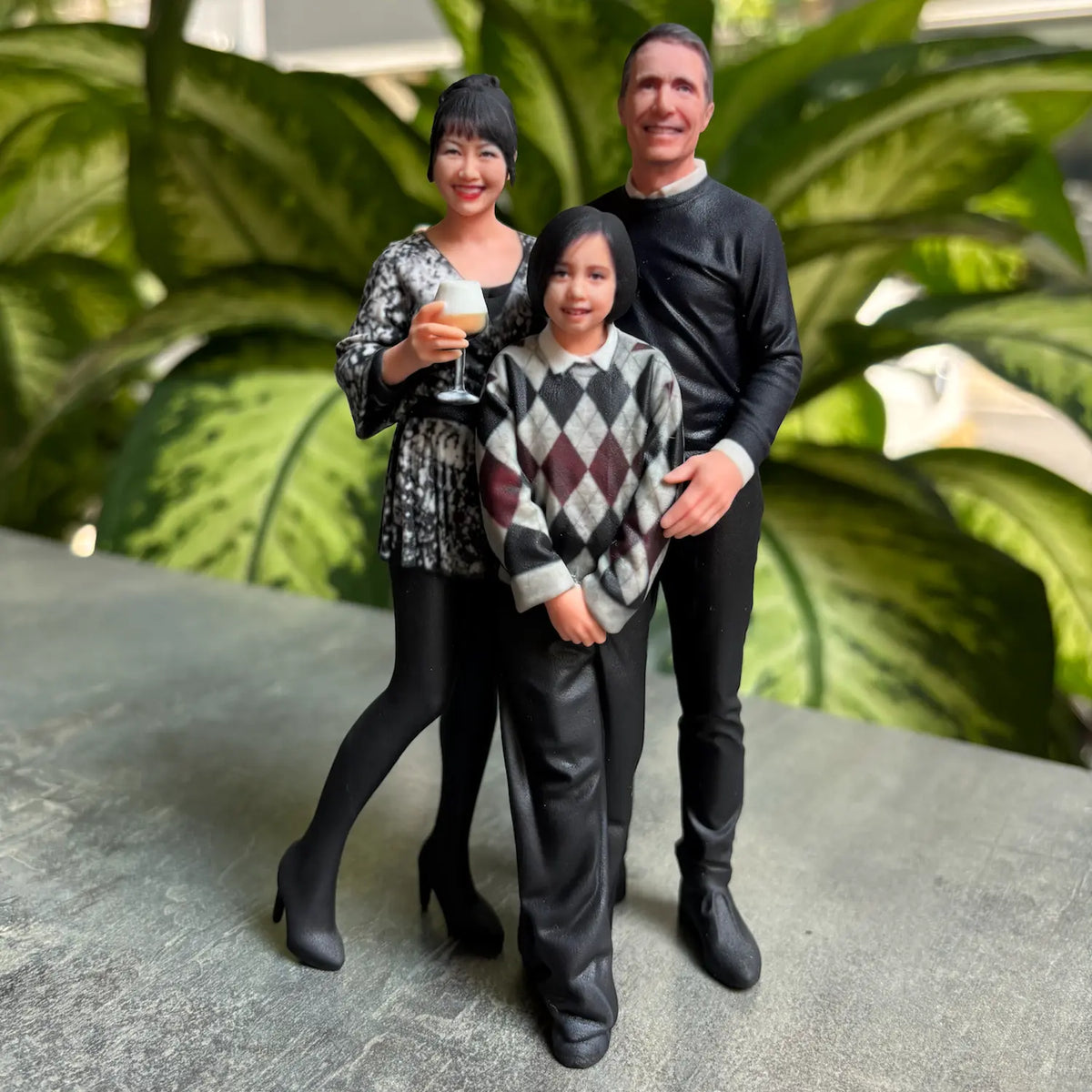 Custom 3D family figurine of three — mother with a champagne glass, father in black and their daughter in an argyle sweater, displayed on a stone surface — 3D Portrait Lab